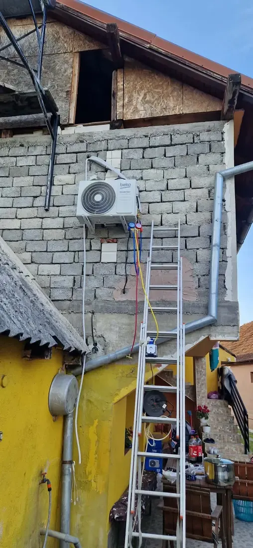 Unitate exterioară de aer condiționat montată pe un zid de cărămidă, cu o scară rezemată dedesubt și furtunuri colorate legate la ea