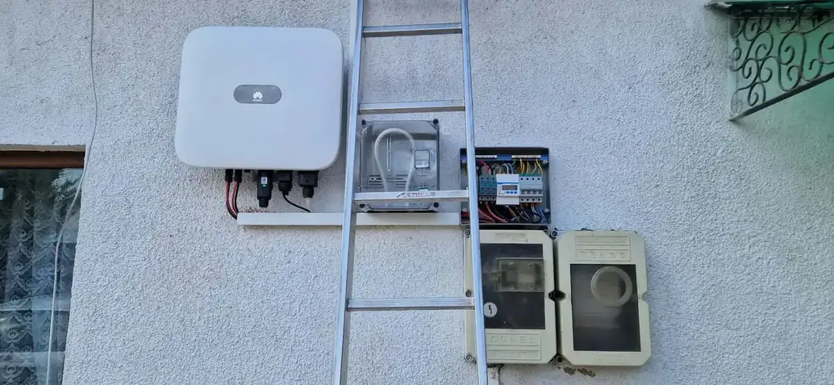 Invertor alb și un tablou electric mic, montate pe peretele unei case, lângă două cutii de plastic bej