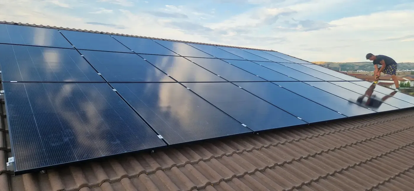 Un om la lucru lângă un șir de panouri fotovoltaice montate pe un acoperiș de țiglă, la apus