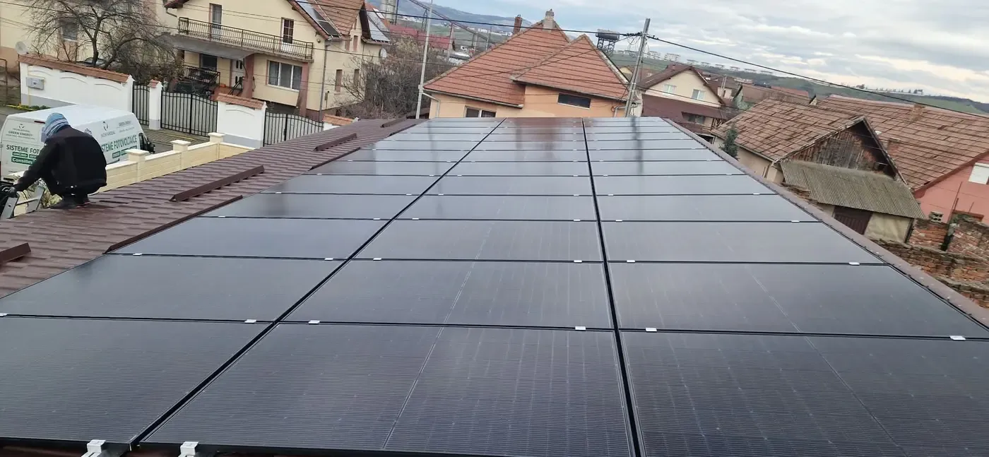 Panouri fotovoltaice negre pe un acoperiș, cu un om la lucru în stânga și case în fundal