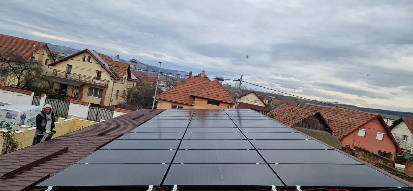 Panouri fotovoltaice negre pe un acoperiș de țiglă maro, cu case și dealuri în fundal și un om la lucru în stânga
