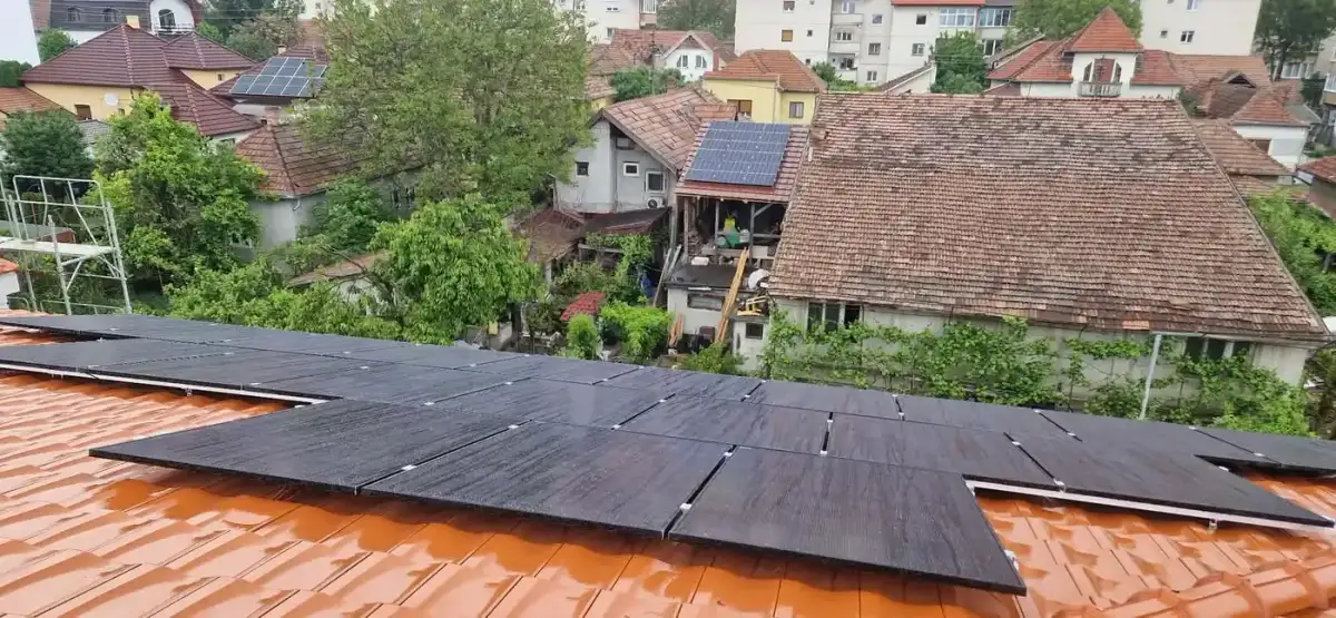 Panouri fotovoltaice negre pe un acoperiș de țiglă portocalie, văzute de sus, cu case în jur