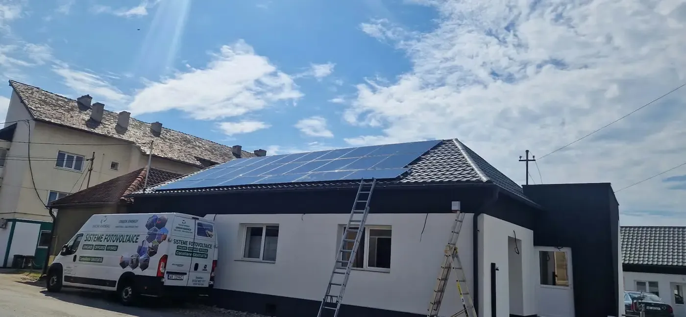 Casă albă cu panouri fotovoltaice pe acoperiș, două scări rezemate de perete și o dubiță albă cu inscripție în față