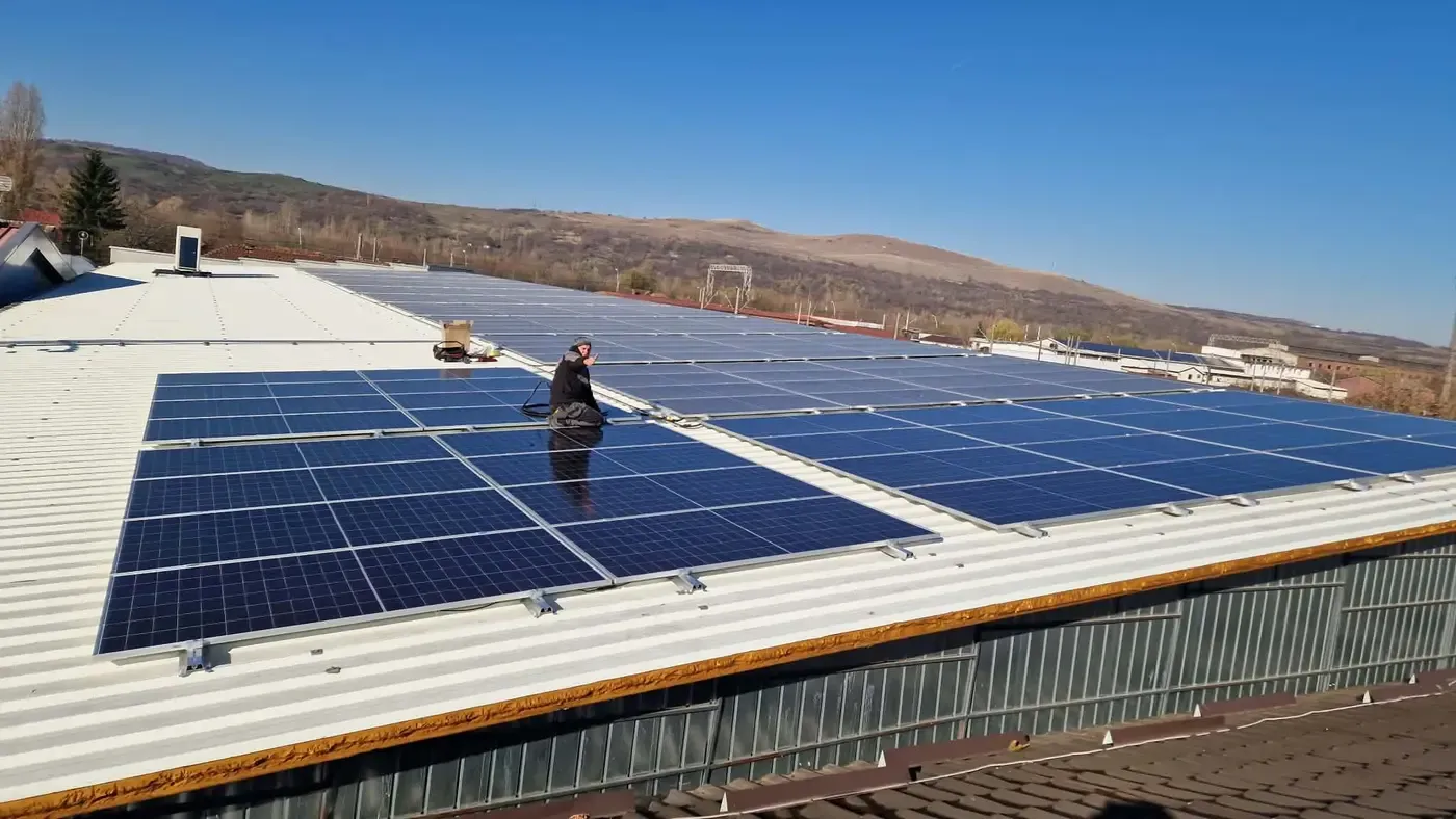 Panouri fotovoltaice pe acoperișul de tablă al unei hale, cu un om la lucru și dealuri în fundal
