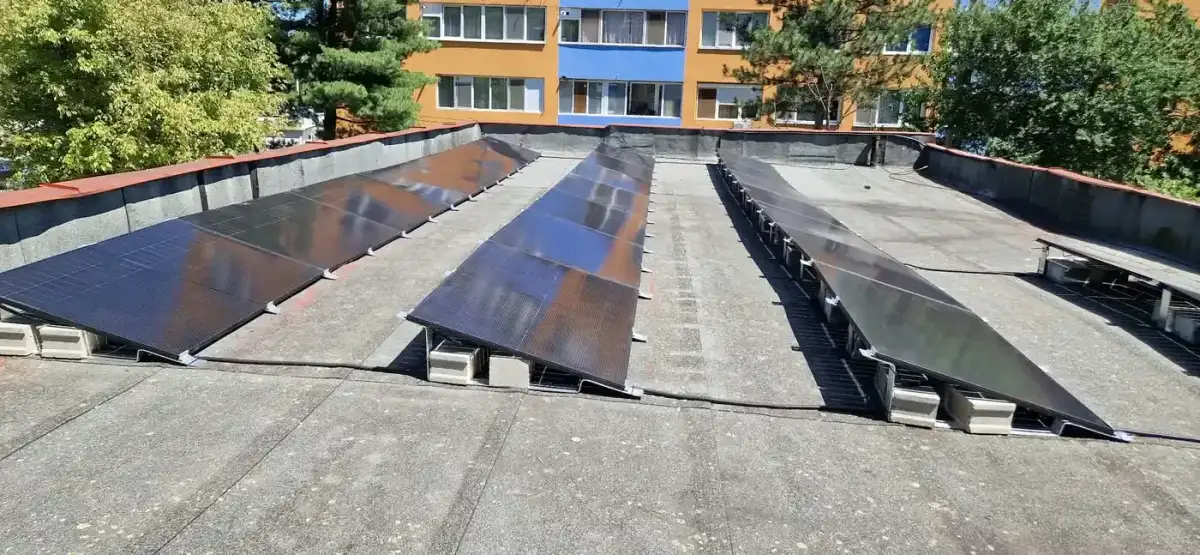 Panouri fotovoltaice înclinate, așezate pe suporți pe o terasă plană, cu blocuri în fundal